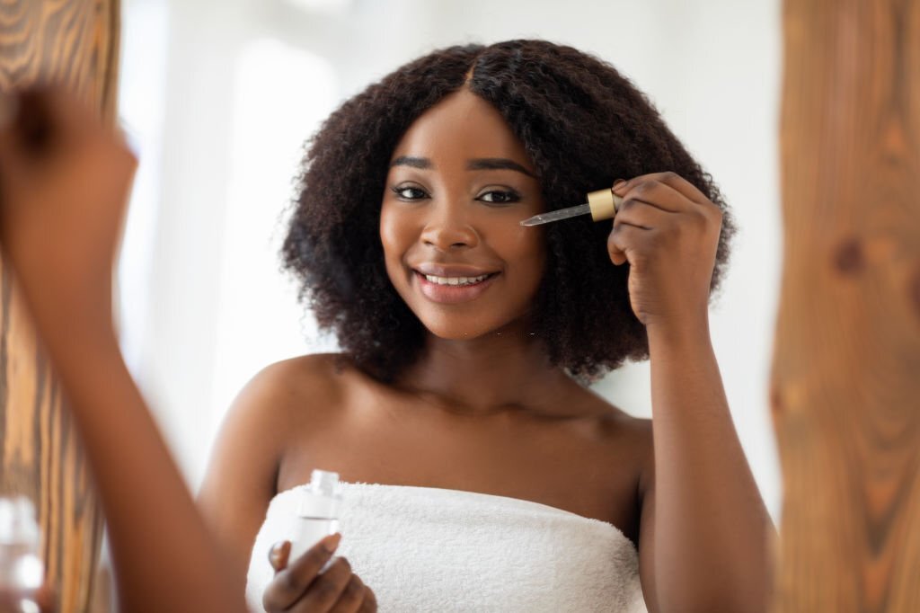 A Smiling young black lady in towel using facial serum near mirror. 